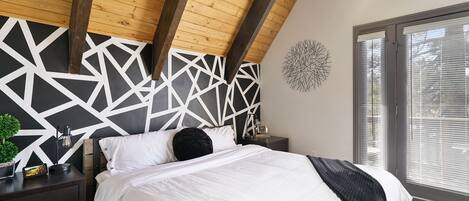 Un dormitorio con techo inclinado, cabecera de diseño geométrico, cama con ropa de cama blanca, mesita de noche con lámpara y ventana con persianas.