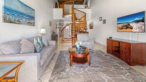 A spacious living room with a spiral staircase, a large painting of a beach, a sofa, a coffee table, and a TV on the wall. - KAMAOLE SANDS 7402, REMODELED, AC, PARTIAL OCEAN, KAMAOLE BEACH PARK! (Kihei)