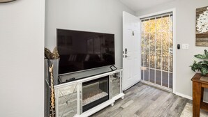 Sala de estar com televisão de tela plana em um rack branco, porta de vidro deslizante para área externa e mesa lateral de madeira com planta.