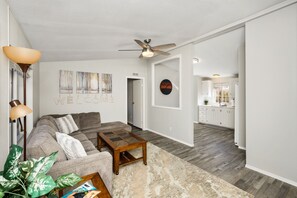 A modern living room with a sofa, coffee table, and a ceiling fan. There is a kitchen visible through an open doorway. - Pet-Friendly. Fully fenced yard (Prescott)