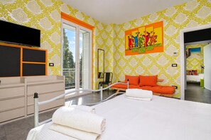 Une chambre d’hôtel moderne avec un grand lit, un coin salon avec des coussins orange, un tableau au mur et une vue sur l’extérieur.