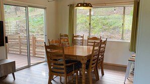 Un espace repas comprenant une table et des chaises en bois, de grandes fenêtres avec des rideaux et un plafond avec des poutres apparentes.