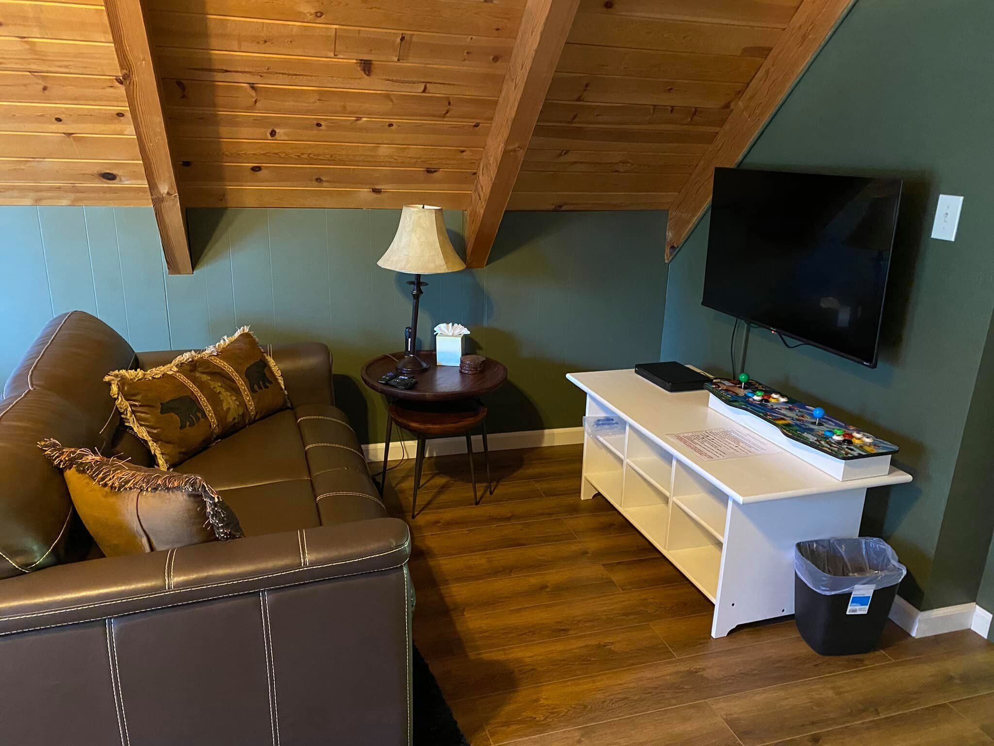 Een woonkamer met een bruine leren bank, een kleine tafel met een lamp, een aan de muur bevestigde televisie en een spelconsole.