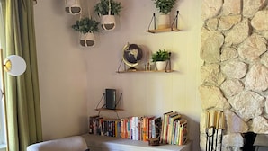 Un salon chaleureux avec un mur en pierre, une cheminée, une bibliothèque et un coin salon confortable comprenant un fauteuil et un pouf.