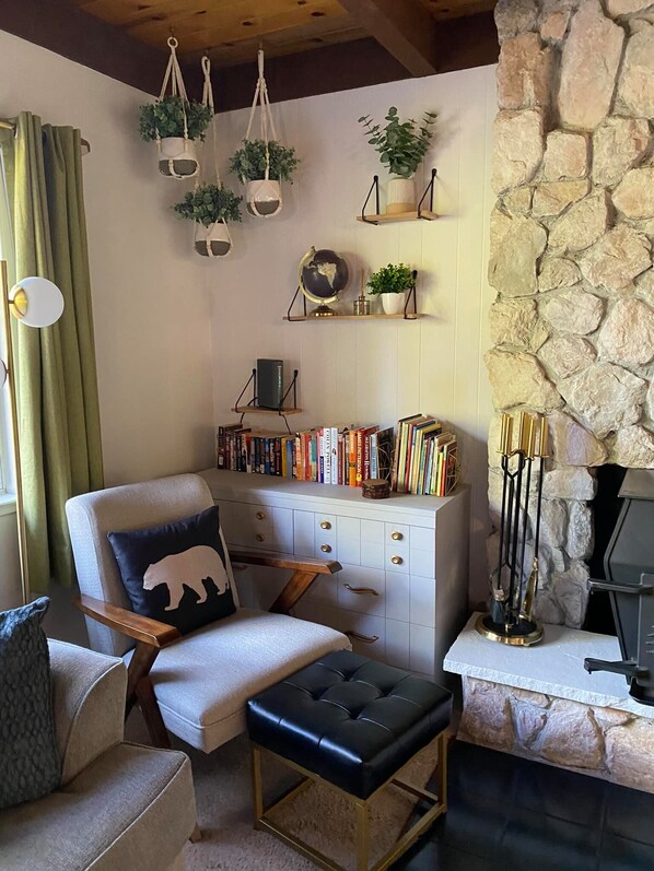 A cozy living room with a stone wall, a fireplace, a bookshelf, and a comfortable seating area with a chair and ottoman.