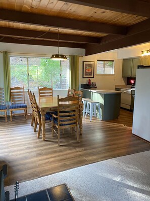 A dining area with a wooden table and chairs, a kitchen with a refrigerator and stove, and a view of greenery through large windows.