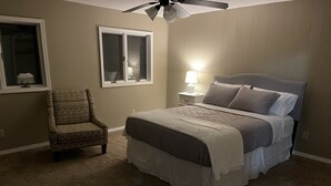 Un dormitorio con cama, una silla, una mesita de noche con lámpara, un ventilador de techo y una ventana.