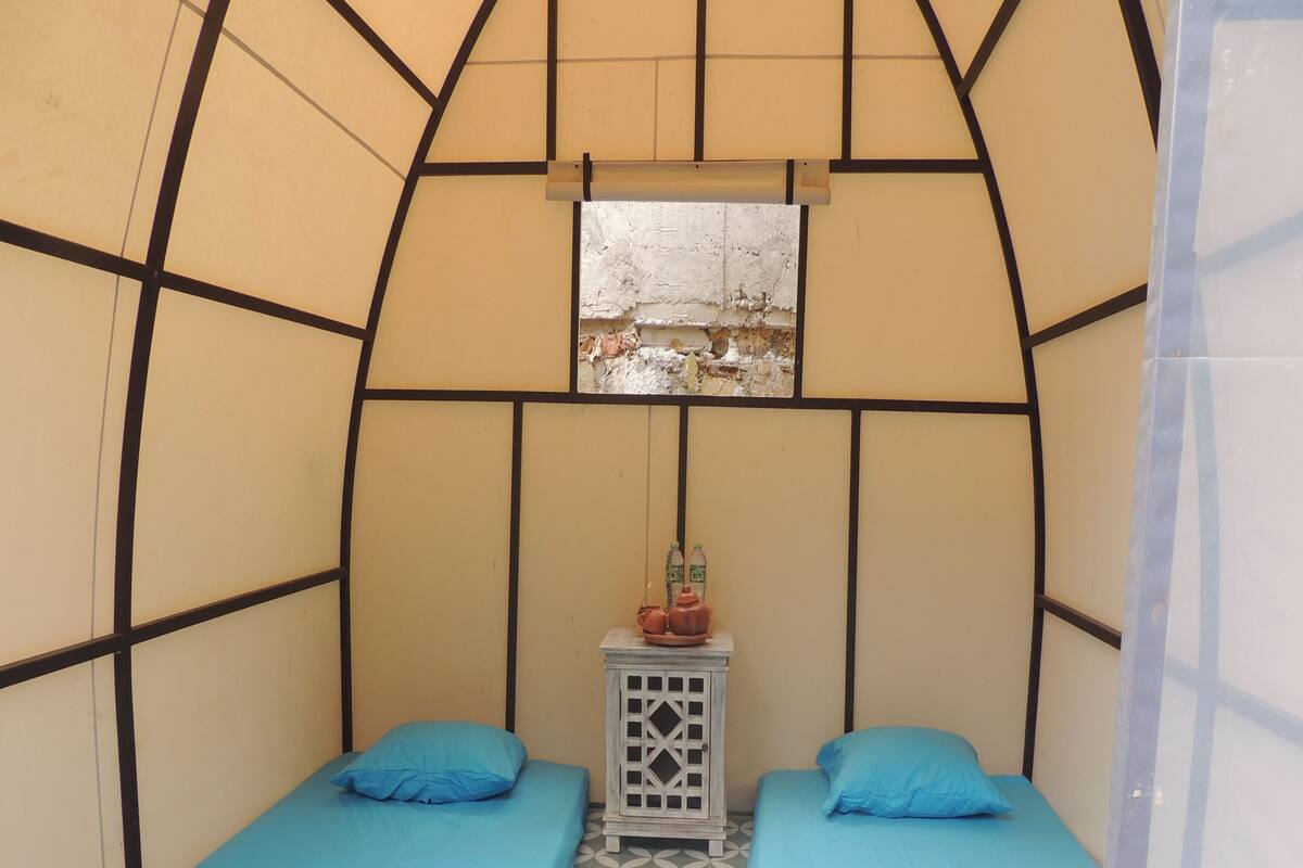 Una habitación pequeña y privada con dos camas, cada una con colchón y almohada azules. Hay una pequeña mesa con una botella de agua y un objeto decorativo encima. Las paredes son beige con detalles en negro, y el suelo tiene un patrón de baldosas geométricas.