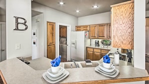 A modern kitchen with wooden cabinets, a refrigerator, and a dining area with plates and napkins. - Bella Views! Biking/Walking Trail System Hub! (Bella Vista)