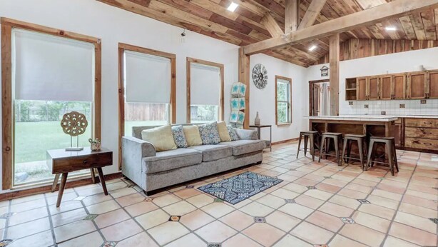 Amplia sala de estar con un sofá gris, vigas de madera en el techo y una cocina con armarios de madera.