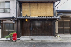 Un bâtiment traditionnel japonais, avec une porte en bois, une petite boîte aux lettres rouge et des plantes en pot disposées sur le trottoir.