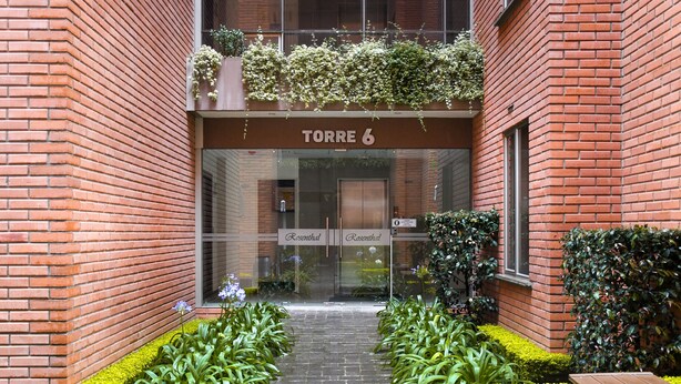 Fachada de edificio de ladrillo, un letrero que dice 'TORRE 6', y un pequeño jardín con bancos y plantas.