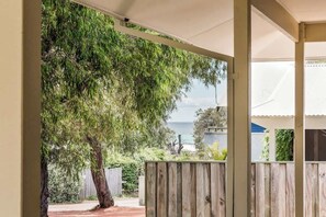 A covered outdoor area with a view of the ocean, a wooden fence, and a tree. - Reverie By The Bay—Walk to Beaches, Relax in Style (Dunsborough)