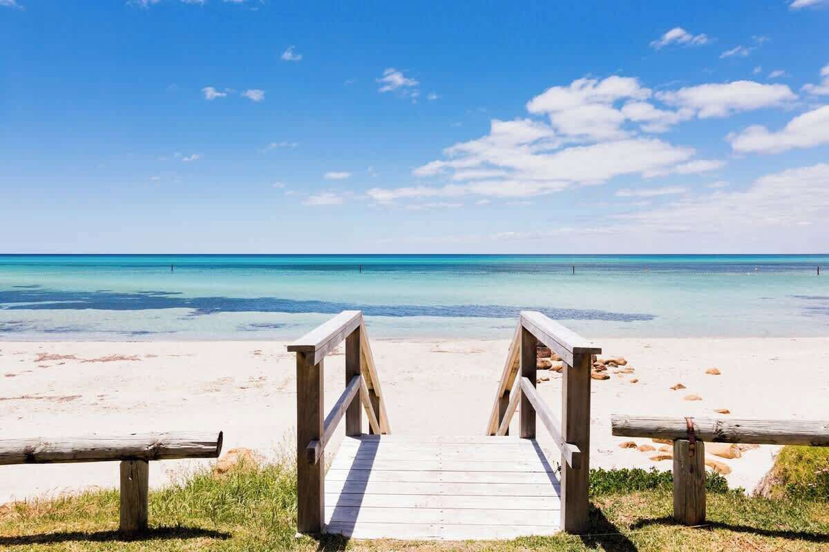 Ein hölzerner Steg führt zu einem Strand mit klarem türkisblauem Wasser und wolkenlosem Himmel.