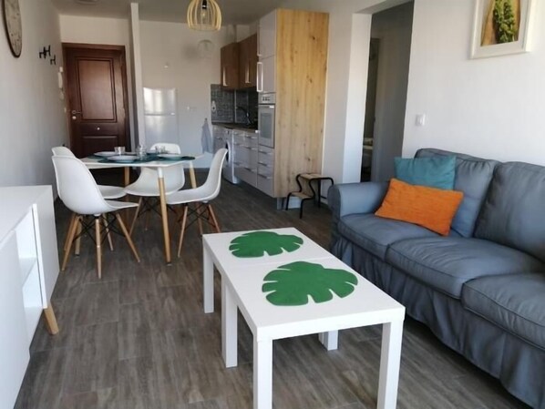 A modern living room with a grey sofa, a white coffee table with a green leaf design, and a dining area with white chairs and a table.