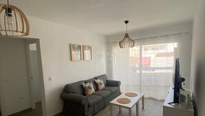 Ein modernes Wohnzimmer mit grauem Sofa, zwei gerahmten Kunstwerken, einem Couchtisch mit Untersetzern, einem Fernseher auf einem Schrank und einer Balkontür.