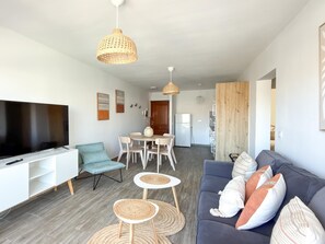 A modern living room with a flat-screen TV, a grey sofa with patterned cushions, a dining area with a wooden table, and a kitchen in the background. - Elcano by Best Holidays Fuerteventura (Corralejo)