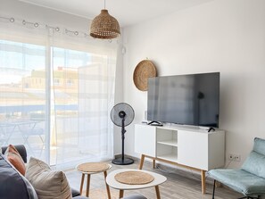 A modern living room with a sofa, a TV on a white cabinet, a fan, and a view of buildings through large windows.