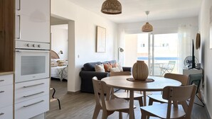 Una cocina moderna con armarios blancos y horno integrado, un comedor con mesa y sillas redondas, y una sala de estar con sofá y un ventanal grande.