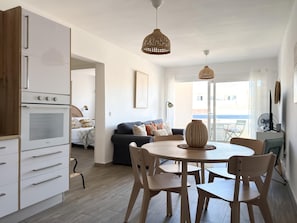 Una cocina moderna con armarios blancos y horno integrado, un comedor con mesa y sillas redondas, y una sala de estar con sofá y un ventanal grande.