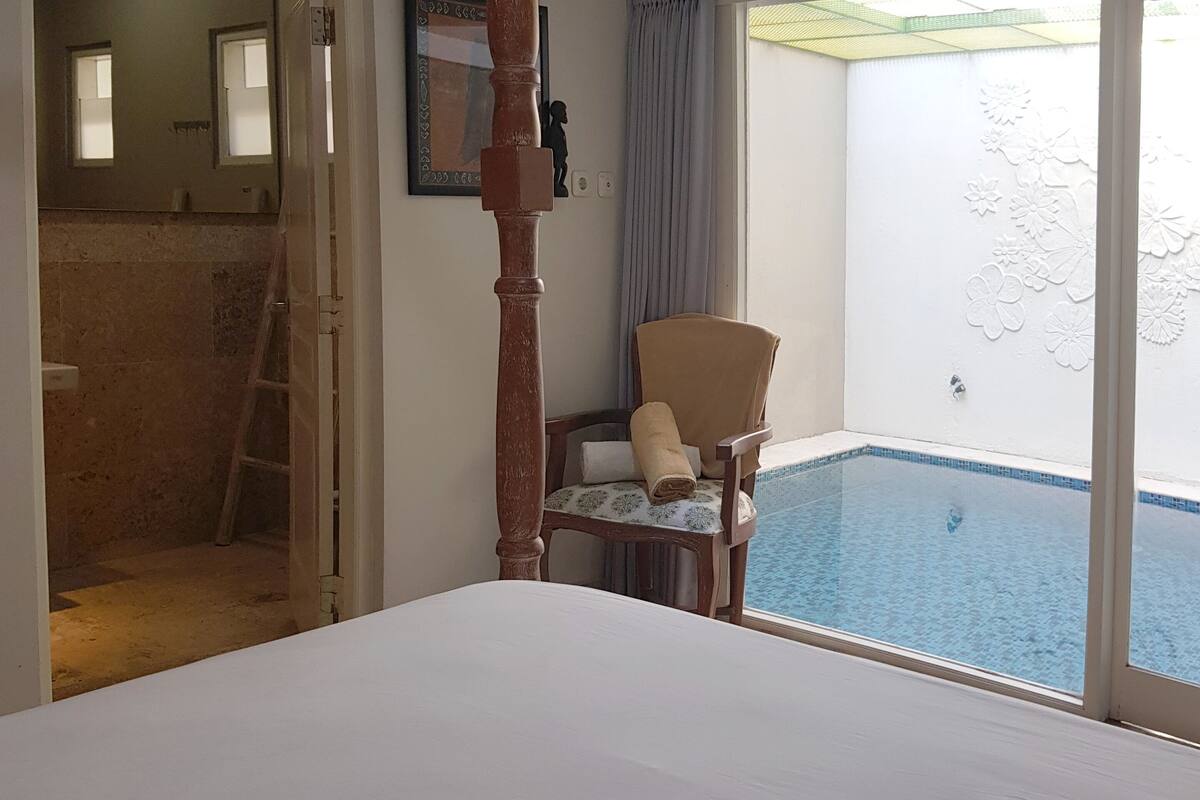 Quarto com cama, uma cadeira com toalha, uma mesa pequena e vista para uma piscina.
