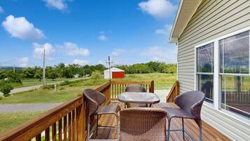 A wooden deck with wicker chairs and a table, overlooking a grassy area with a house and a red building in the distance.