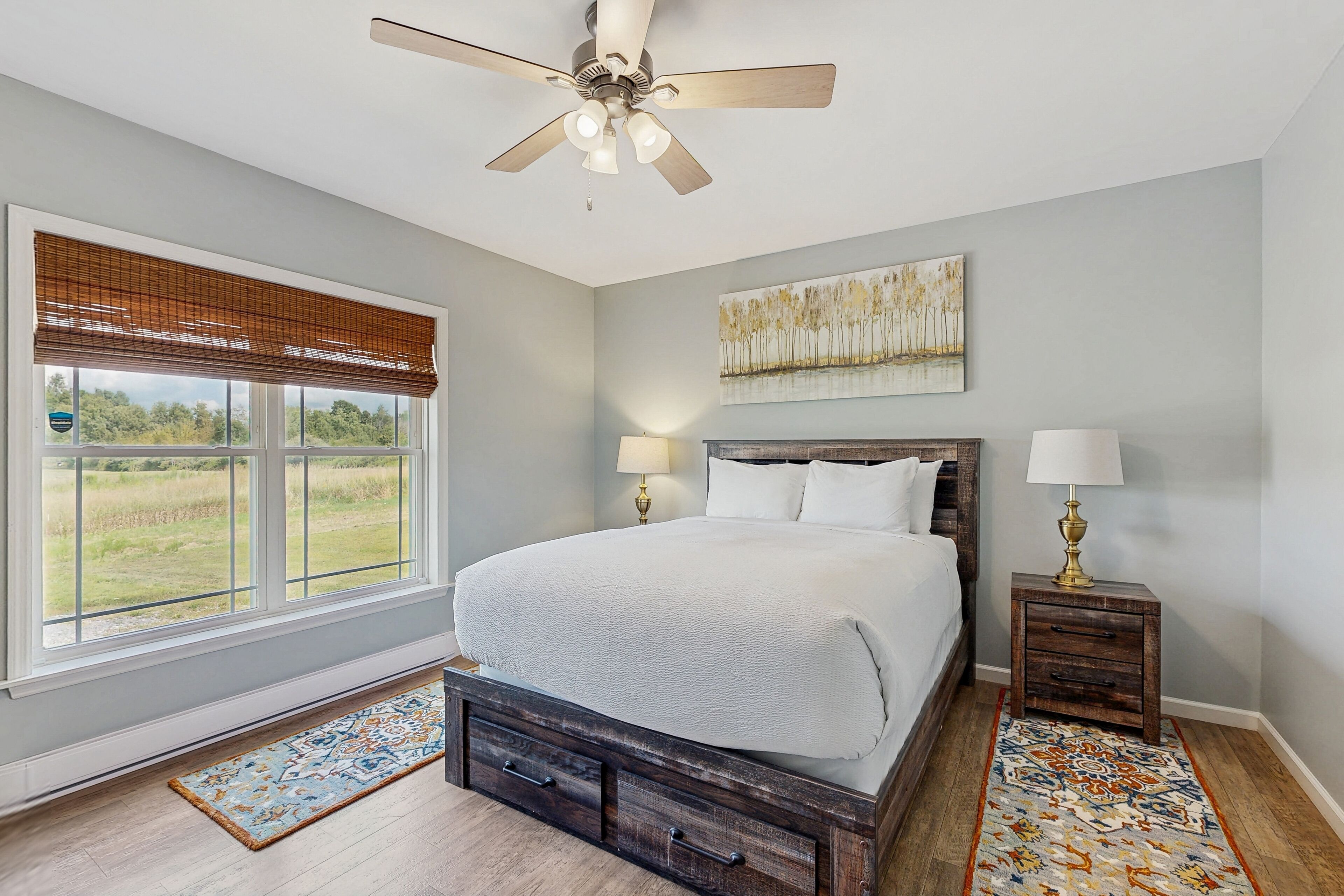 A bedroom with a bed, two bedside tables, a ceiling fan, a window with blinds, a rug, and a painting on the wall.