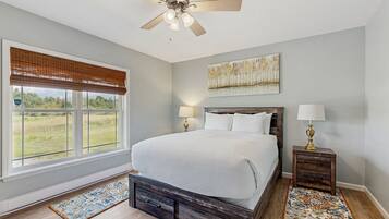 A bedroom with a bed, two bedside tables, a ceiling fan, a window with blinds, a rug, and a painting on the wall.