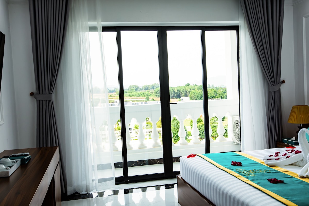 A hotel room with a balcony, a bed with a teal bedspread, a desk with a lamp, and a view of greenery outside.