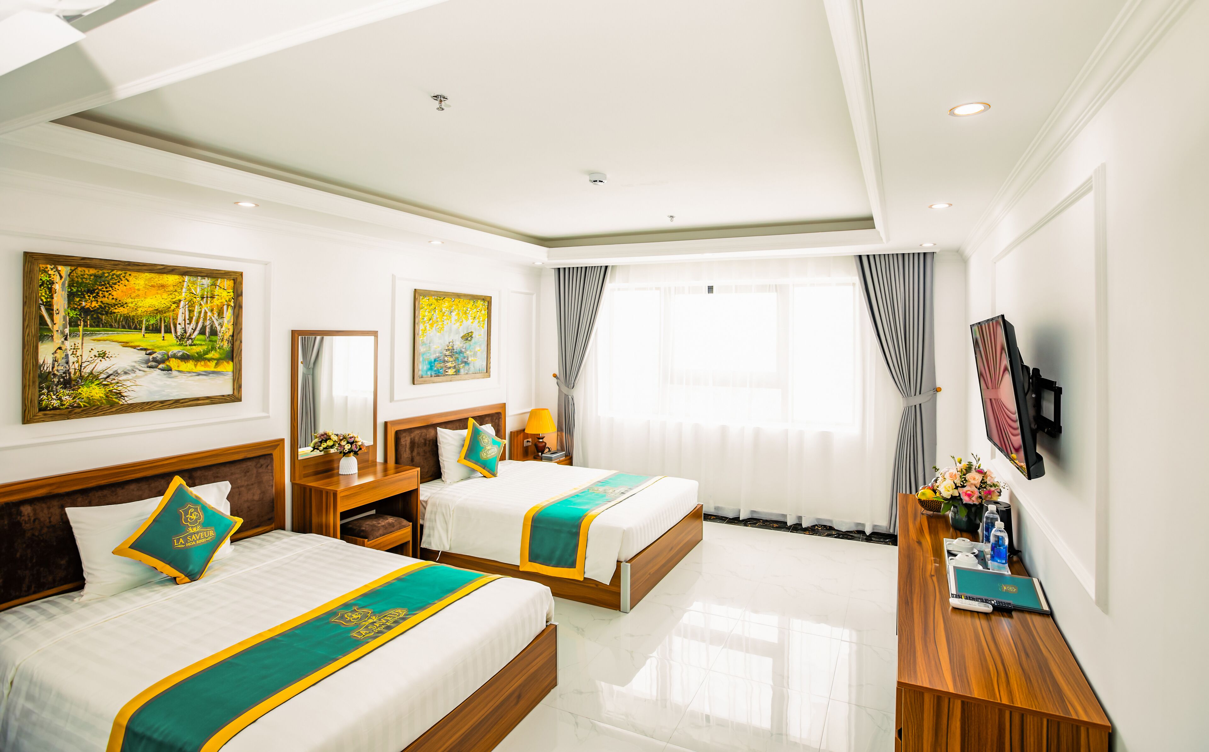 A hotel room with two beds, a desk, a TV, and artwork on the walls.