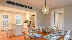 Un comedor con una mesa de madera preparada para seis comensales, sillas blancas y una cocina al fondo.