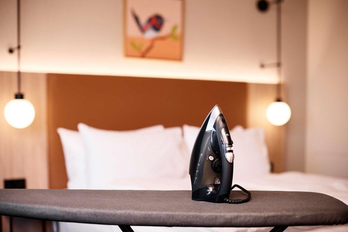 A neatly made bed with white linens, a wall-mounted painting of a bird, and two pendant lights.
