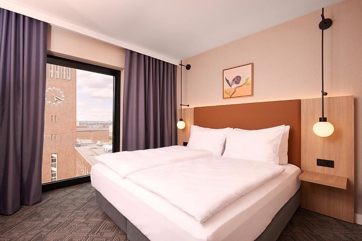 Habitación de hotel con una cama grande, vista panorámica de la ciudad a través de la ventana y un reloj en la pared.