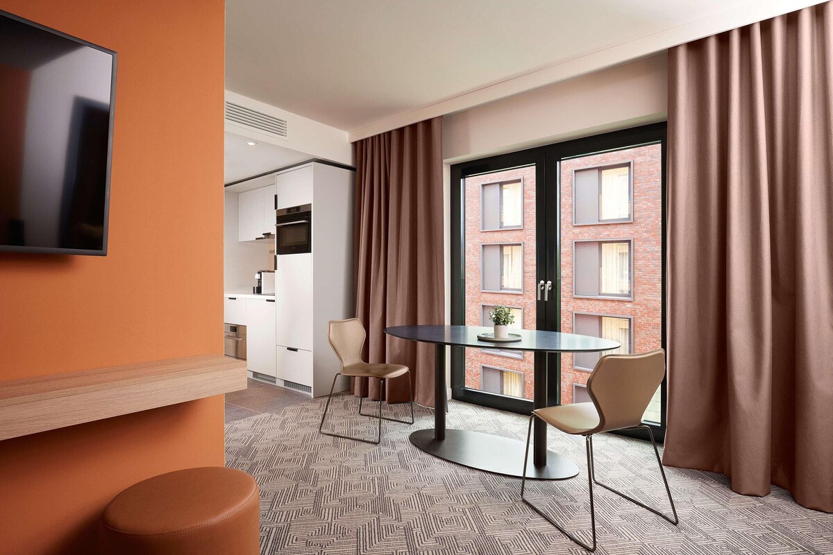 Una habitación de hotel moderna con una mesa redonda, dos sillas, un televisor montado en la pared y un ventanal con cortinas.