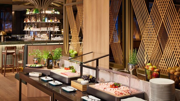 A buffet setup with various food items and utensils, a bar area with shelves of bottles, and a wooden decorative wall.