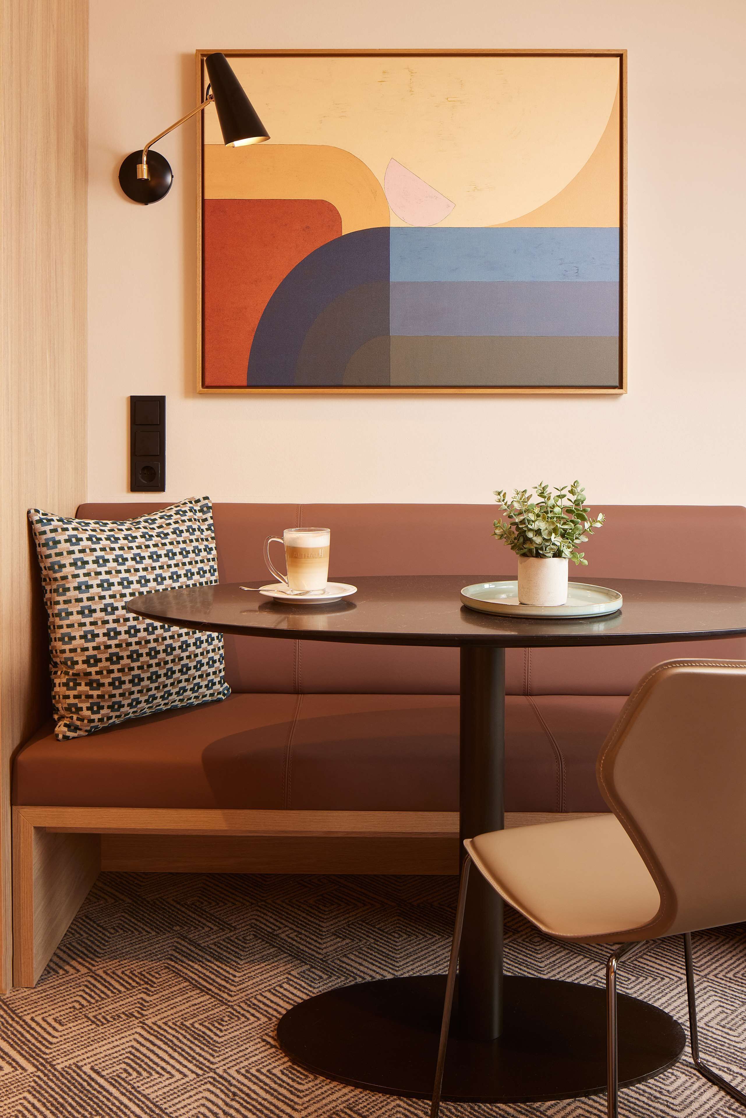 Un área de estar moderna con una mesa redonda, un banco acolchado, un cojín estampado, una pequeña planta en maceta y una taza de café.