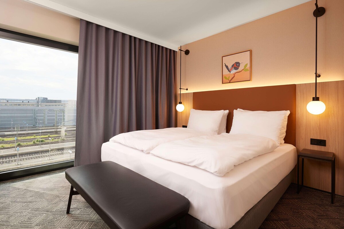 Ein Hotelzimmer mit einem großen Bett, einer Bank, einem Nachttisch und Blick auf die Stadt durch große Fenster.