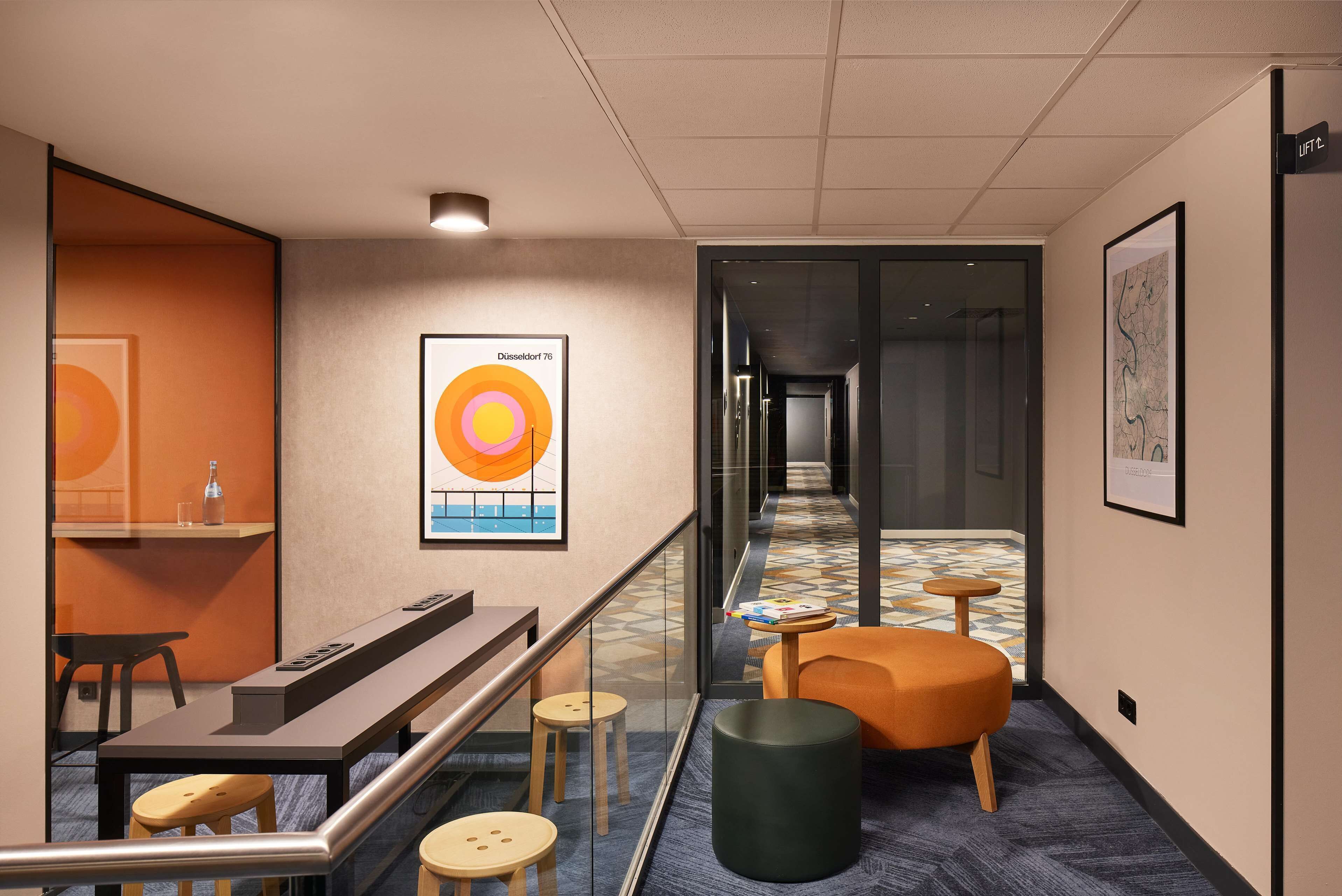 A modern hotel lobby with a long reception desk, a seating area with stools and a cushioned chair, and a patterned floor.