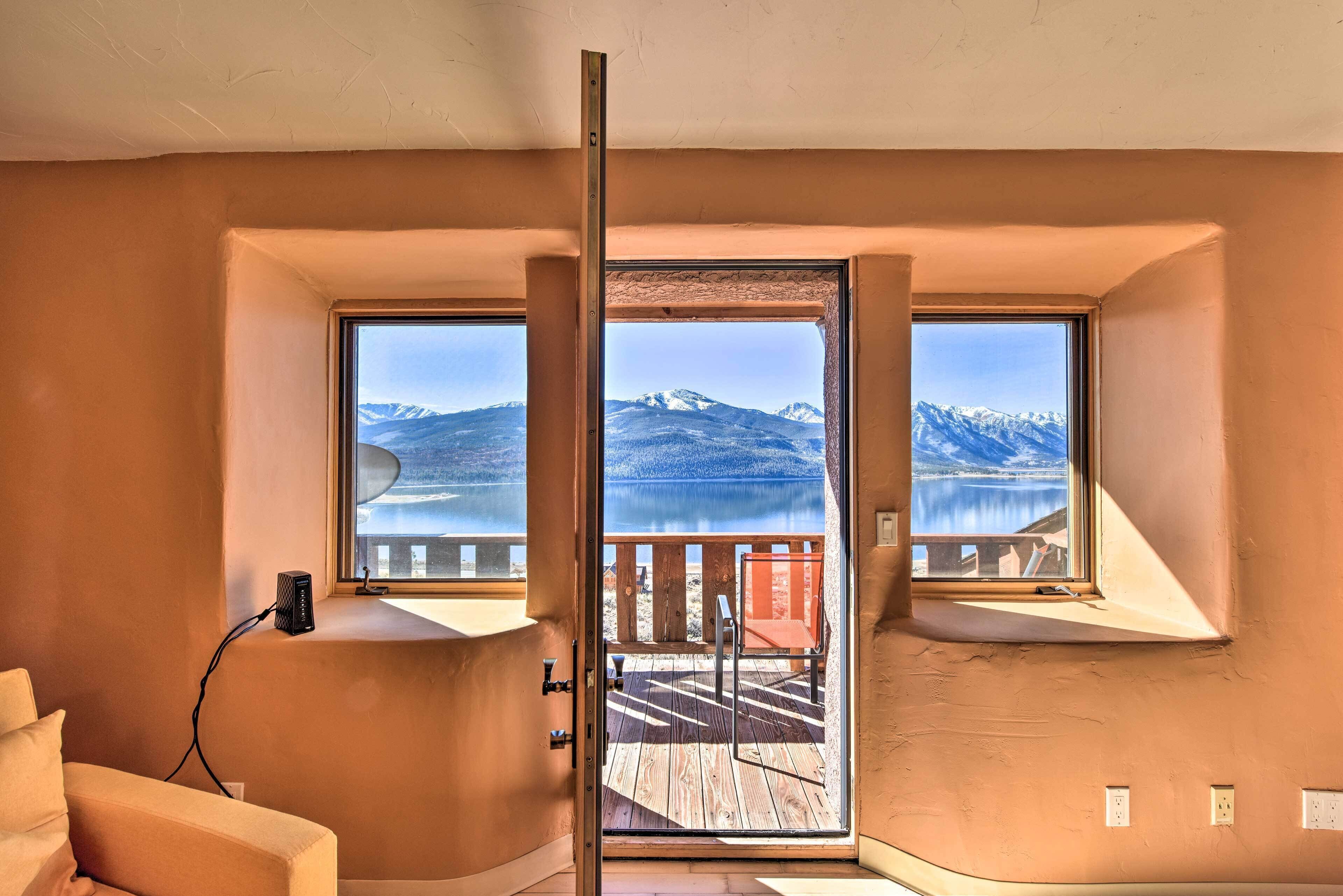 A room with a large window overlooking a lake and mountains. There is a wooden deck outside the window.