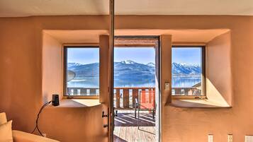 A room with a large window overlooking a lake and mountains. There is a wooden deck outside the window.