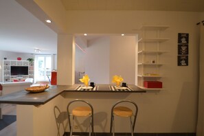 Una cucina moderna con isola centrale, due sgabelli da bar e un'area pranzo con TV e scaffali.