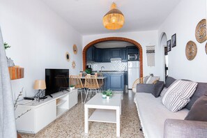 Una sala de estar con sofá, televisor y mesa de centro, una cocina con gabinetes azules y un arco de ladrillo, y un comedor con sillas y una mesa.