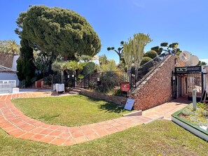 Un jardín ajardinado con muro de piedra, un camino empedrado y diversos árboles, incluyendo un conífero grande y un cactus.