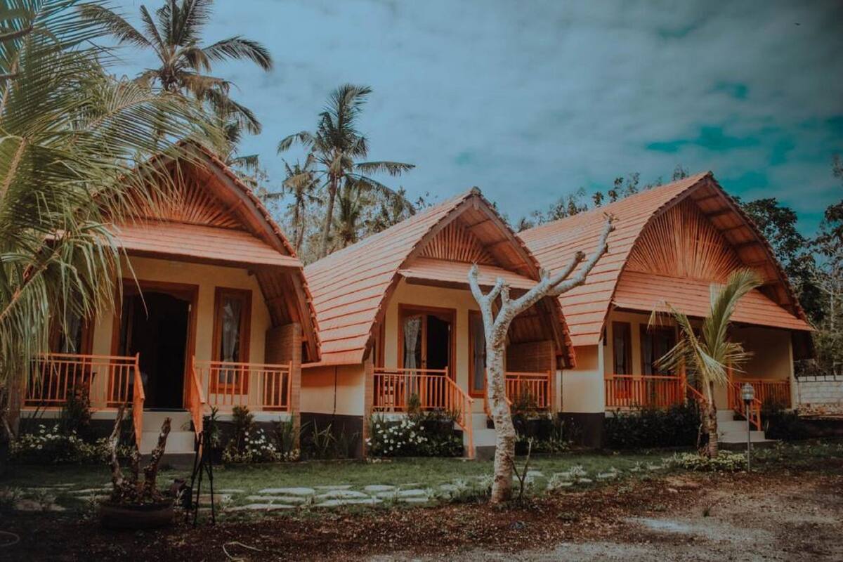 Een rij van drie vrijstaande huisjes in bungalowstijl, met oranje daken en houten balkons, omringd door palmbomen en een heldere lucht.