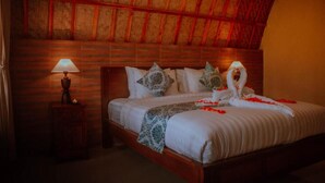 A room with a wooden ceiling, a bed with white bedding, multiple pillows, a bedside lamp, and a decorative arrangement of flowers and a swan on the bed. - Watu Jineng by WizZeLa (Penida Island)