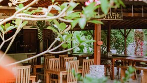 An outdoor wooden dining area with tables and chairs, surrounded by lush greenery and a covered structure. - Watu Jineng by WizZeLa (Penida Island)