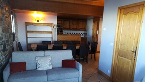 A living room with a grey sofa, a dining table with chairs, a kitchen in the background, and a wooden door.