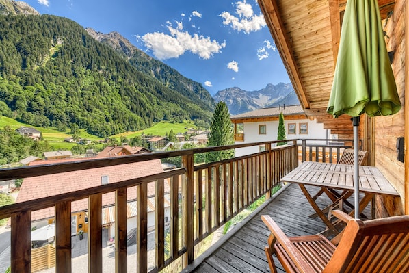 A wooden balcony with a table and chairs, overlooking a mountainous landscape and a village below.