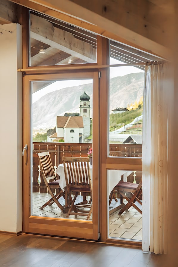 Una habitación con puerta corrediza de vidrio que da a una terraza exterior con muebles de madera y vistas a una montaña e iglesia.