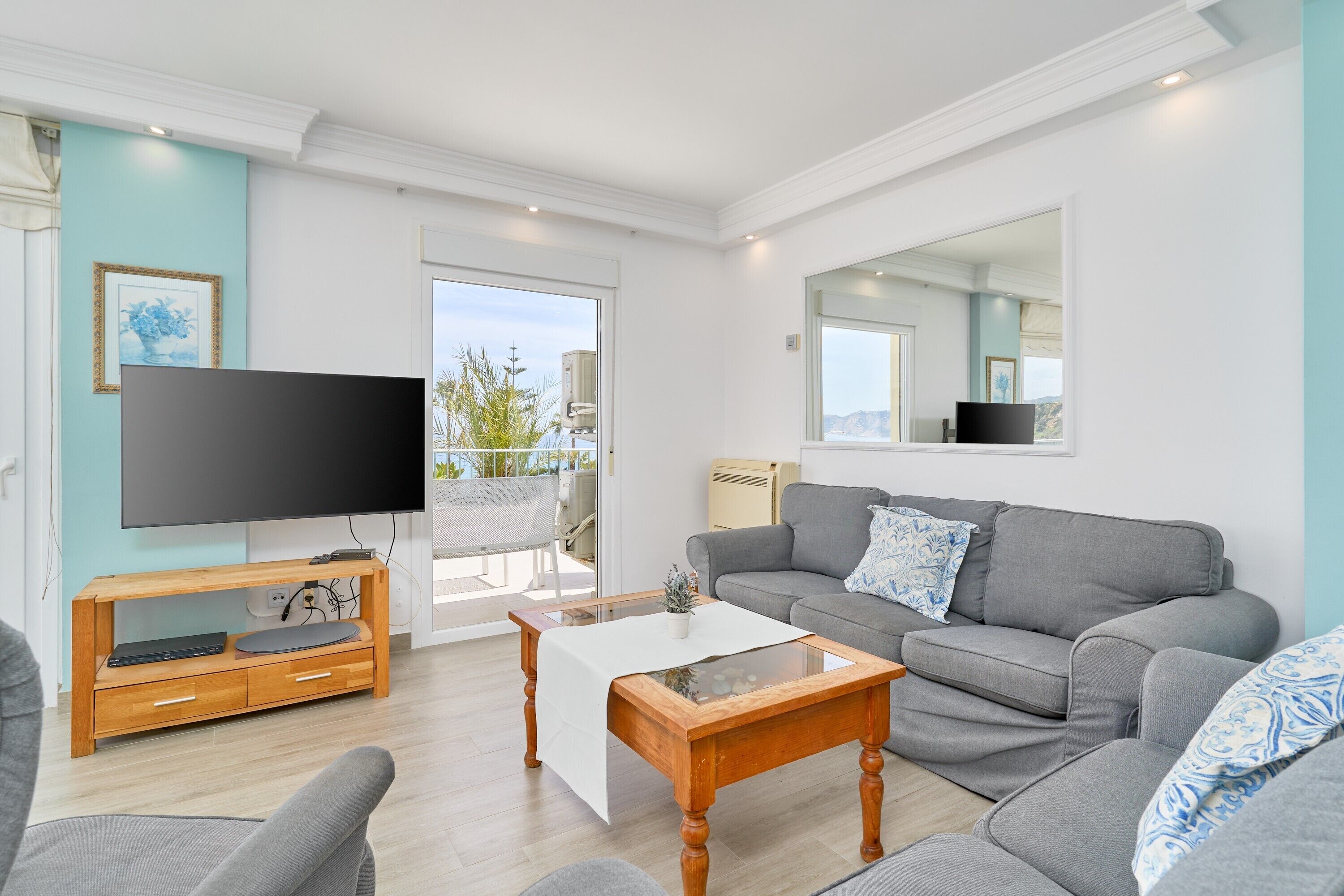 A living room with a grey sofa, a wooden coffee table, a flat-screen TV on a wooden stand, and a balcony with a view.